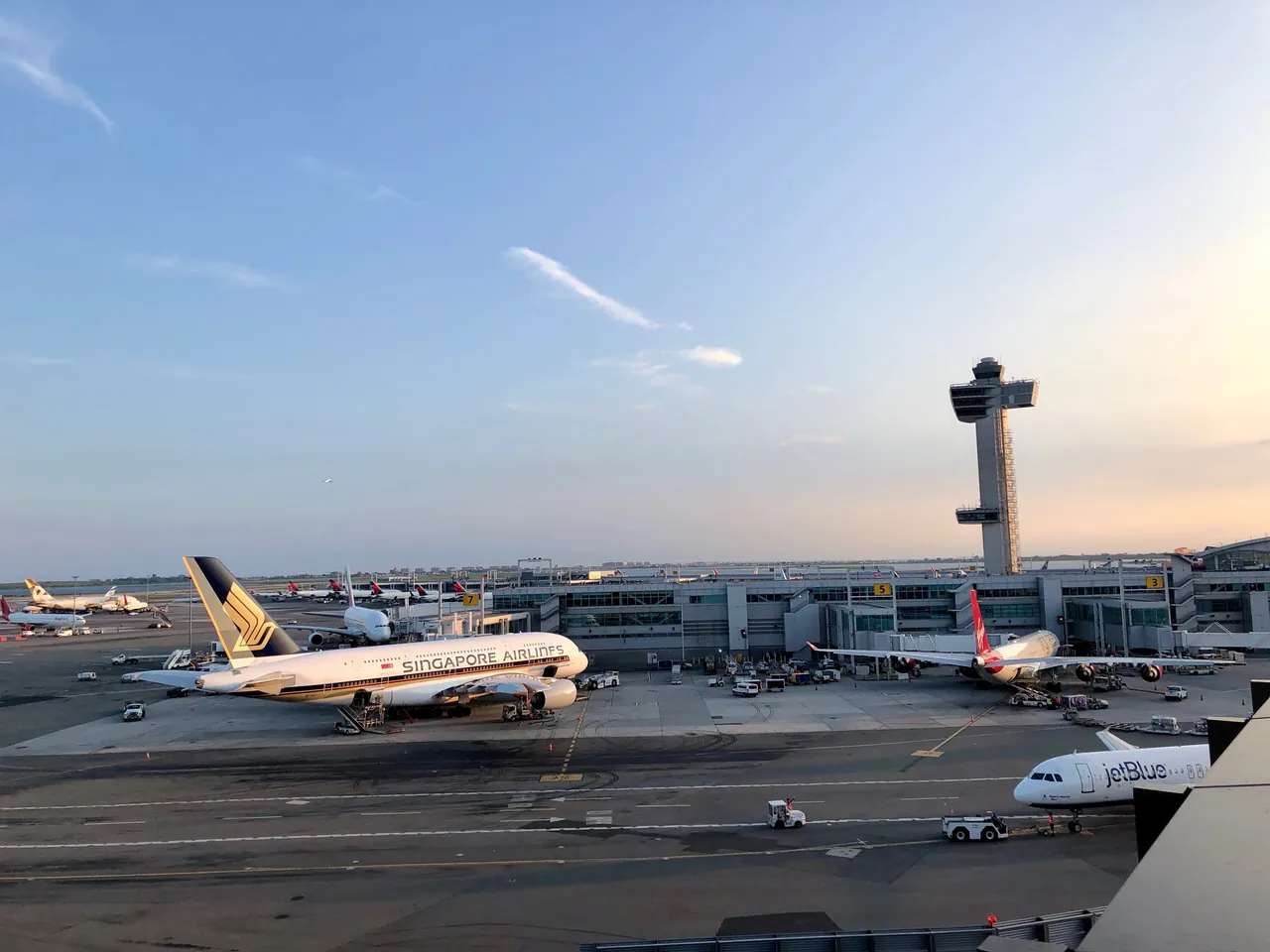 Singapore Airlines A380 and a Virgin Atlantic jet at their gates at JFK Terminal 4 at sunset, with the control tower rising behind the terminal